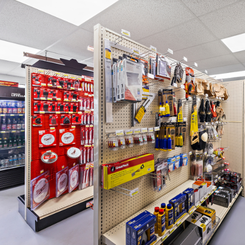 Three aisles: third aisle shows a refrigerator with beverages, second aisle has Milwaukee saw blades and accessories, first aisle contains tool belts, knee pads, and some hand tools like a hand saw and screwdriver.