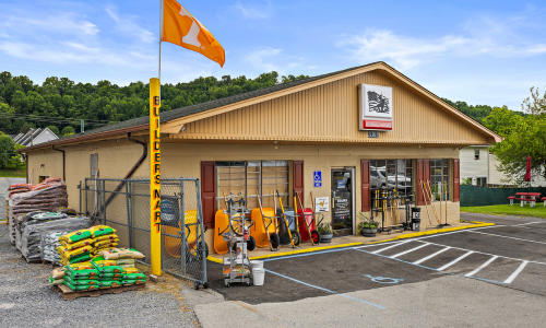 Storefront of BLOUNTVILLE HARDWARE. Beige building with an orange University of Tennessee flag. Lawn and garden supplies on the left, wheelbarrows by the door, and tools like rakes and pickaxes on the right.