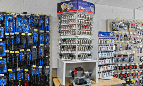 Our Ilco keys selection, featuring silver, gold, and black-capped keys, sits on top of the key cutting station. To the right are keys with patterns like an owl, American flag, and orange camo. To the left is a display of Century Drill & Tool products, and to the right are wire wheels.